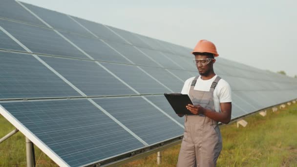 Técnico de pie con portapapeles en la estación solar. Hombre africano en mono, casco naranja y gafas de seguridad de pie en la estación solar y la escritura en el portapapeles. Concepto de mantenimiento y personas. — Vídeos de Stock