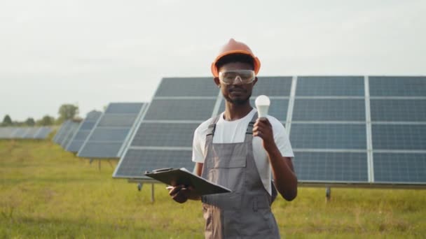 非洲裔美国人手持太阳能电池板和灯泡站在战场上。穿着棕色工作服、橙色头盔和安全眼镜的男性技师。关于绿色能源的想法。太阳能农场 — 图库视频影像
