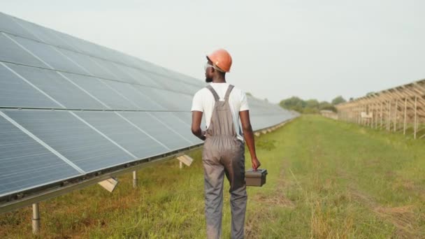 Vue arrière de l'homme afro-américain en uniforme et casque à l'aide d'instruments tout en travaillant sur le terrain avec des panneaux solaires. Production d'énergie alternative. Ingénieur avec instruments marchant sur la ferme solaire — Video