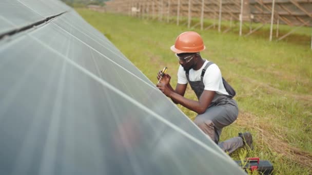 Üniformalı ve kasklı bir adam istasyondaki güneş panellerini tamir etmek için tornavida kullanıyor. Fotovoltaik hücrelerin kurulumu. Yeşil enerji konsepti. Teknisyen dışarıda tornavidayla güneş panellerini tamir ediyor. — Stok video