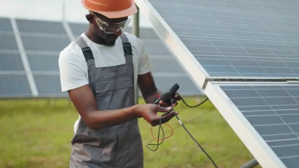 Technicien en uniforme utilisant le multimètre tout en vérifiant la tension dans les panneaux solaires. Afro-américain contrôlant la production d'énergie verte sur la station. Homme en salopette mesurant la résistance dans les panneaux solaires — Video
