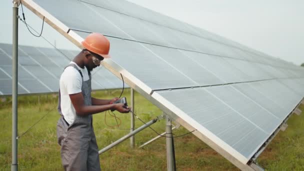 Ingeniero africano profesional en casco y uniforme utilizando multímetro para medir el amperaje en paneles solares. Concepto de personas, mantenimiento y energía alternativa. Hombre en overol midiendo resistencia — Vídeos de Stock