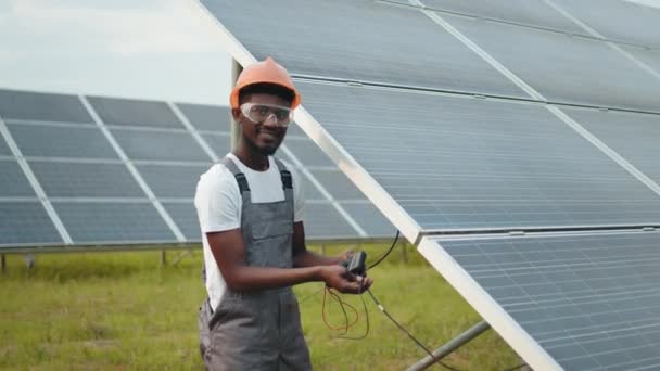 Инженер измеряет напряжение в солнечных батареях на открытом воздухе. Африканский человек в сером комбинезоне измеряет сопротивление солнечных панелей на открытом воздухе. Компетентный техник, использующий мультиметр во время осмотра на станции. — стоковое видео