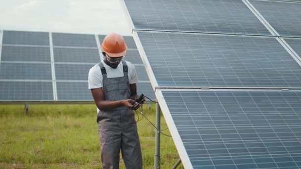 Afroamericani che controllano la produzione di energia verde in stazione. Uomo in tuta grigia misura la resistenza nei pannelli solari all'aperto. Concetto di persone, manutenzione ed energie alternative. — Video Stock