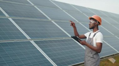 Endüstriyel İşçi güneş istasyonunda panoya yazı yazıyor. Afrika kökenli Amerikalı mühendis, istasyondaki güneş panellerini incelerken panoya notlar yazıyor. Güneş çiftliğinde gri tulumlu bir adam.
