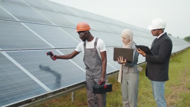 Femme musulmane et homme indien debout avec un technicien africain et vérifier la température des panneaux solaires. Inspecteurs utilisant ordinateur portable et presse-papiers, travailleur tenant imageur thermique. Inspection de la station solaire — Video