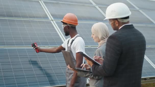 Muslimische Frau mit Laptop und indischer Mann mit Klemmbrett stehen mit afrikanisch-amerikanischem Techniker auf einer Solarfarm. Industriearbeiter zeigt Inspektoren Wärmebildkamera mit Indizes. — Stockvideo