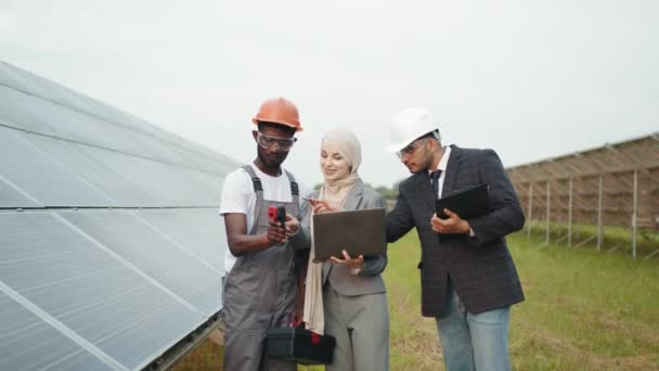 Pracovník v průmyslu ukazuje inspektorům termokamery s indexy. Africký americký technik ukazující teplotu solárních panelů na termokamerách muslimské ženě v hidžábu a indiánskému muži v helmě — Stock video