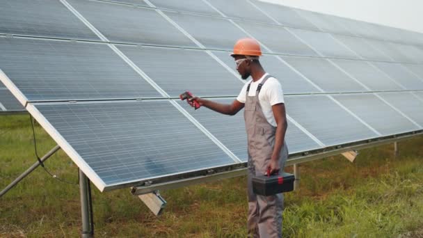 Ingénieur afro-américain en uniforme, casque de sécurité et lunettes utilisant un imageur thermique pour mesurer la chaleur thermique des panneaux solaires. Homme travaillant sur l'équipement de maintenance sur la station moderne. — Video