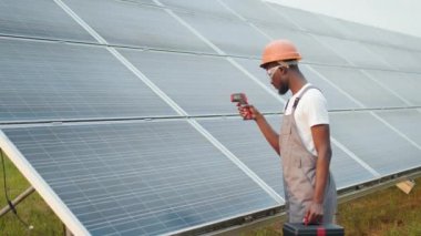 Güneş panellerinin ısısını ölçmek için termal görüntüleyici kullanan güvenlik kaskı mühendisi. Modern istasyonda bakım ekipmanları üzerinde çalışan bir adam. Güneş panellerinin sıcaklığını ölçen mühendis