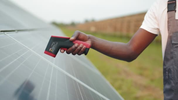 Technik v helmě pomocí termokamery na sluneční farmě. Boční pohled na Afroameričana stojícího venku a měřícího teplo solárních panelů termokamerou. Profesionální technik solární farmy — Stock video