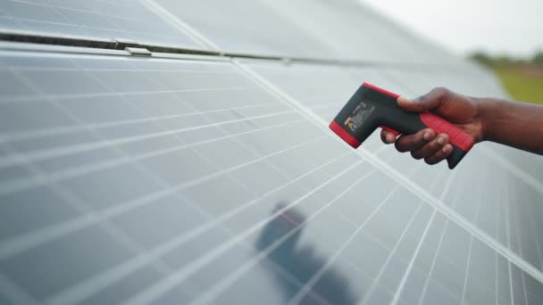 Zbliżenie męskiej dłoni trzymającej elektroniczny termowizor w pobliżu ogniwa słonecznego na zewnątrz. Technik w kasku używając termowizora na farmie słonecznej. Inżynier w jednolitej temperaturze pomiarowej paneli słonecznych — Wideo stockowe