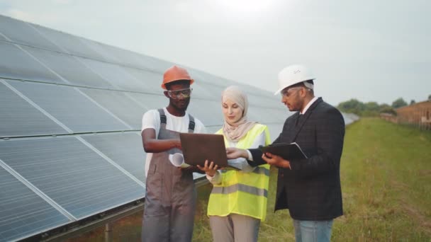 Muslim woman in hijab and indian man in white helmet talking with african american technician on solar station. Multiracial people using laptop, clipboard and blueprints for work outdoors. — Stock Video