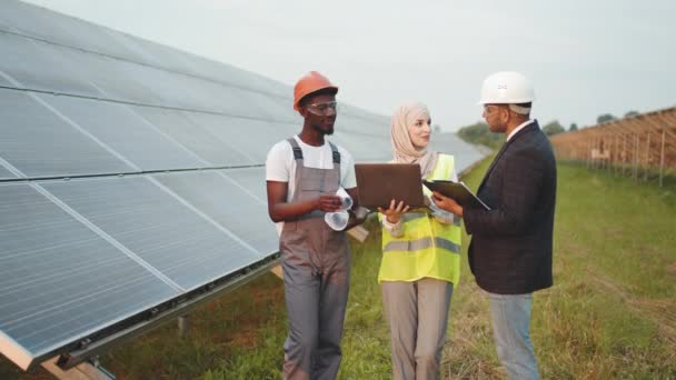 Técnico afroamericano reuniéndose con dos inspectores multirraciales en la estación solar. Hombre de negocios indio en casco blanco y mujer musulmana en equipo de comprobación de hijab de la granja. En la granja solar — Vídeos de Stock