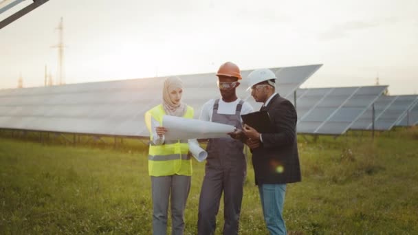 Técnico afroamericano, mujer musulmana y hombre indio de pie juntos en la granja solar y mirando los planos. Las personas multirraciales planean la estrategia de producción de energía alternativa. Estación solar — Vídeos de Stock