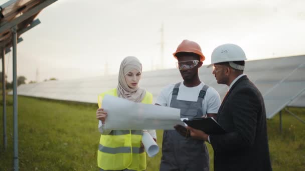 Oameni multirasiali care stau cu planuri pe staţia solară. Grupul de trei oameni multirasiale în căști de siguranță care au întâlnire de lucru între stația solară. Tehnician cu ingineri pe stația solară — Videoclip de stoc