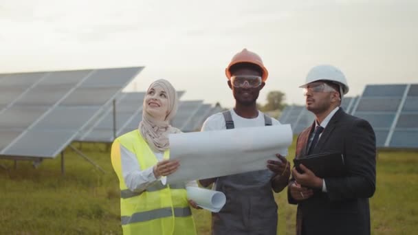 Competent engineers discussing details of project. Ecology and green energy concept. Engineers standing outdoors with blueprints in hands. Three multiracial people having meeting among solar station — Stock Video