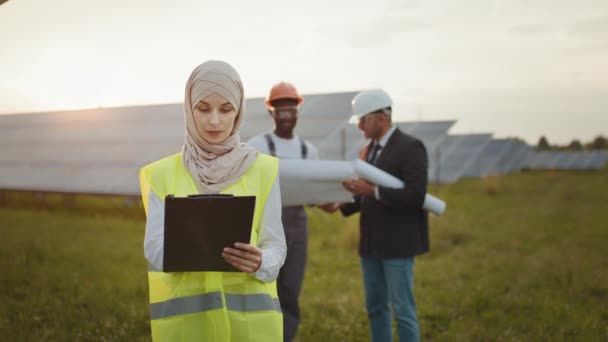 Woman in hijab looking at camera and writing on clipboard while standing on field with solar panels. African american technician and indian engineer having discussion on background. — Stock Video