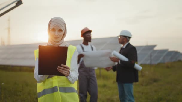 Mujer en hijab usando portapapeles para trabajar. Mujer en hijab mirando a la cámara mientras está de pie en el campo con paneles solares. Dos hombres multiculturales en cascos parados detrás con el plan de proyecto en las manos. — Vídeo de stock