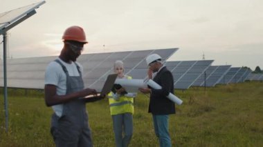 Üniformalı Afro-Amerikan teknisyen güneş panelleri ile sahada duruyor ve dizüstü bilgisayarda daktilo yazarken Müslüman kadın ve Hintli adam arkasında planlarla çalışıyor. Alternatif enerji kavramı.
