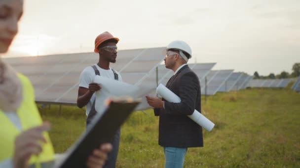 Side view of muslim woman in hijab writing on clipboard while standing among rows of solar panels. Two multicultural men in helmets standing behind with project plan in hands. — Stock Video