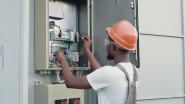 Üniformalı ve kasklı Afrikalı elektrikçi elinde kablolarla kutunun yanında duruyor ve güneş istasyonunda servis yapıyor. Bakım, insanlar ve yeşil enerji konsepti. Güneş çiftliğindeki teknisyen