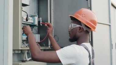 Turuncu kasklı ve güvenlik gözlüklü Afrikalı Amerikalı tamirci güneş istasyonu arasındaki güç transformatöründeki kabloları kontrol ediyor. Yeşil enerji üretiminde uzman teknisyen denetim süreci.