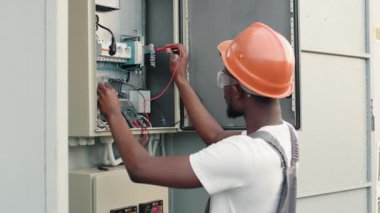 Tamirci güneş çiftliğindeki güç transformatörünün kablolarını kontrol ediyor. Güneş istasyonu arasında turuncu kasklı Afrikalı Amerikalı tamirci. Yeşil enerji üretiminde uzman teknisyen denetim süreci.