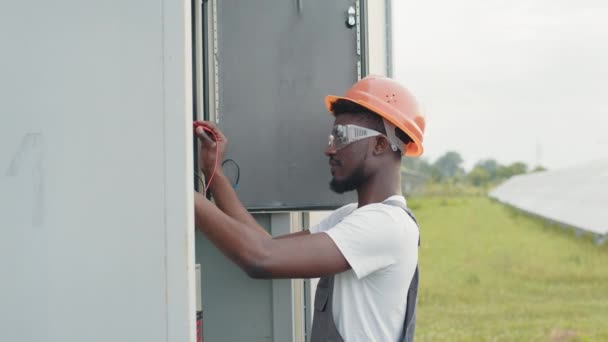 Tecnico competente in casco arancione e tuta grigia in piedi vicino agli interruttori e alla stazione solare di connessione alla rete comune. Produzione di energia alternativa e risparmio ambientale. — Video Stock
