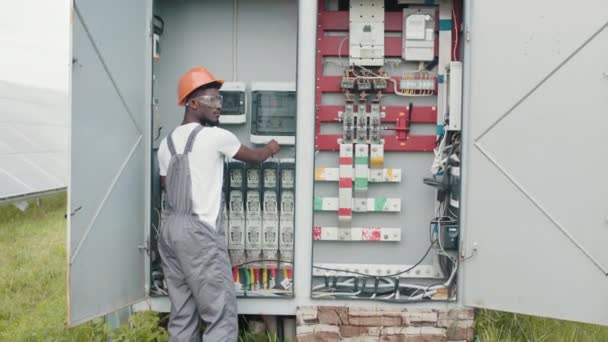 Hombre afroamericano en mono gris y casco naranja reparando cables en aparamenta en la estación solar. Concepto de personas, mantenimiento y energía alternativa. — Vídeo de stock