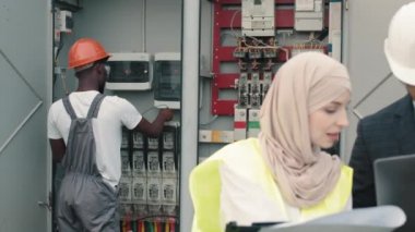 Güç transformatörü. Elektrik Sistemleri Güç Dağıtım Kabinesi. Çok ırklı insanlar yeşil enerji üretim sürecini inceliyor. Şalter arkasında duran Afrikalı Amerikalı teknisyen