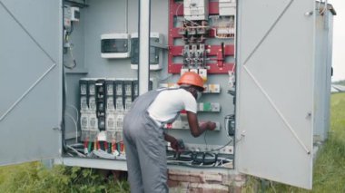 Tulumlu, güvenlik kasklı, gözlüklü, gülümseyen ve güneş istasyonunda şalter başında kameraya bakan Afro-Amerikan bir adam. Alternatif enerji üretimi. Güneş istasyonunda teknisyen