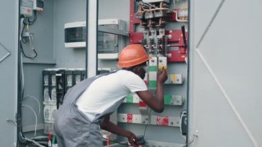 Tulumlu Afro-Amerikan bir adam güneş istasyonunda şalter başında duruyordu. Elektrik Sistemleri Güç Dağıtım Kabinesi. Mühendis Testi Voltaj Akım Elektrik Hattını Ölçüyor.