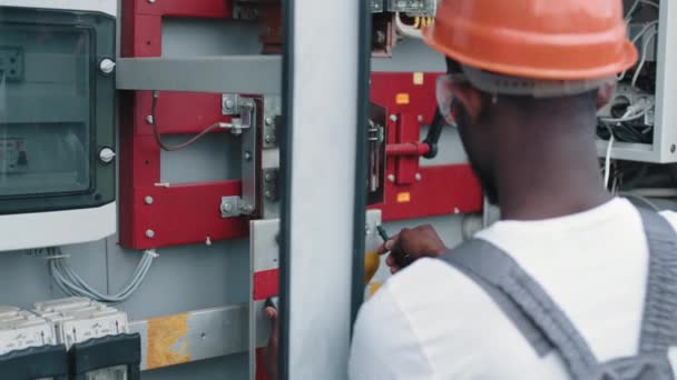 Detailní záběr člověka, který používá multimetr pro práci na solární stanici. Afričtí američtí elektrikáři v bezpečnostní helmě a kontrolním napětí v rozvaděčích při práci na solární stanici. — Stock video