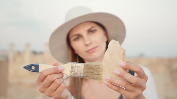 专业考古学家拿着古老的柱子，用刷子擦干净。戴着夏帽和休闲装的年轻女子在户外工作。古代妇女考古发掘 — 图库视频影像