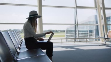 Havaalanında oturan iş kadını büyük bir pencerenin arkasında kucağında dizüstü bilgisayarla oturuyor, işini bitiriyor ve pencereye yaklaşıyor. Freelancer, ofisten uzaktan çalışma konsepti.