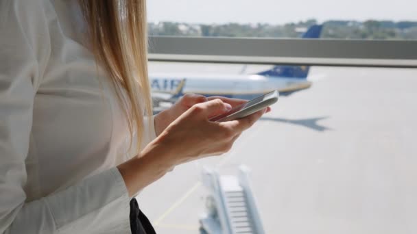ใกล้ชิดของผู้หญิงมือส่งข้อความในผู้ส่งสารหรือส่งข้อความบนสมาร์ทโฟนที่หน้าต่างสนามบิน รูปทรงของมือผู้หญิงที่มีโทรศัพท์ ผู้หญิงเดินทางใช้สมาร์ทโฟนที่สนามบิน . — วีดีโอสต็อก
