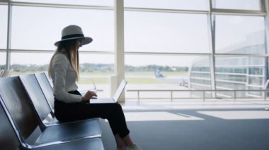 Havaalanında dizüstü bilgisayarda oturan genç ve güzel bir kadın. Kız turist serbest çalışıyor ve bekleme odasında uçmayı bekliyor. Konsept seyahat, uzaktan çalışma. Büyük bir arka planda siluet