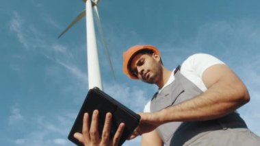 Miğferli ve gri tulumlu Hintli mühendis elinde modern tabletle yel değirmeni çiftliğinin ortasında duruyordu. Foreman büyük türbinlerin çalışmalarını inceliyor ve kontrol ediyor. Yel değirmeni çiftliğinde duran mühendis