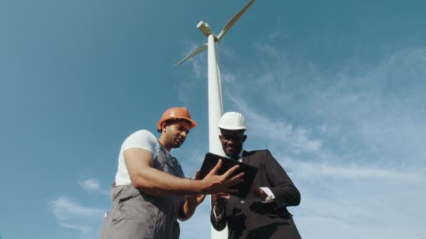 風車農場のエンジニアと検査官が協力しています。スタイリッシュなスーツでアフリカ系アメリカ人検査官と会う際にタブレットを使用して均一でヘルメットでインドのエンジニア. — ストック動画