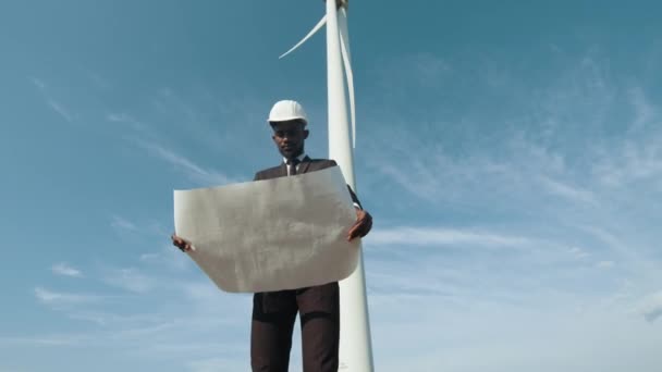Låg vinkel syn på fokuserad afrikansk amerikansk man står på fältet med vindkraftverk och undersöka ritningar som håller i händerna. Begreppet förnybar och hållbar energi. Man med ritningar — Stockvideo