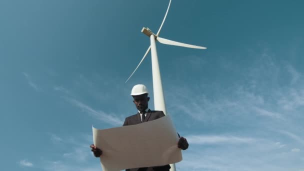 Homme en costume debout parmi les moulins à vent avec des plans. Homme d'affaires afro-américain avec casque blanc tenant des plans et regardant de côté parmi la ferme éolienne. Vue en angle bas. Concept d'énergie verte. — Video