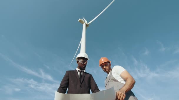 Equipe de empresários e engenheiros em um traje preto a pé explora futuros projetos de instalação de turbinas eólicas. Energia renovável. Inspecção de um projecto num campo de turbinas eólicas que produz electricidade. — Vídeo de Stock