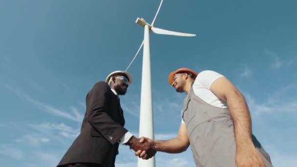 Afrikaanse inspecteur in witte helm en zwart pak schudden handen met indiaanse ingenieur terwijl staan in de buurt van enorme windmolen. Twee partners ingenieurs schudden elkaar de hand na het sluiten van een goed windmolenpark. — Stockvideo