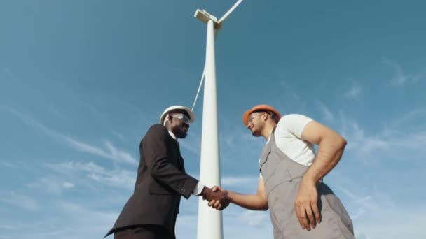 Inspetor africano em capacete branco e terno preto apertando as mãos com engenheiro indiano enquanto estava perto de um enorme moinho de vento. Conceito: energia renovável, tecnologia, eletricidade, serviço, verde, futuro. — Vídeo de Stock