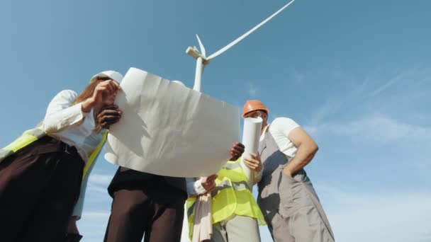 Čtyři multietničtí partneři v bezpečnostních helmách studují plány větrných turbín. Inženýr Diskuse o větrné turbíny Týmová práce a překračování prostředí pole. Ekologická technologie pro elektrickou energii. — Stock video