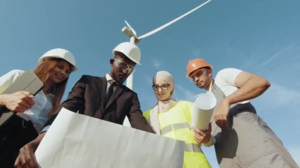 Quatro parceiros multi étnicos em capacetes de segurança estudando plantas de turbinas eólicas enquanto. Engenheiros exploram futuros projetos de instalação de turbinas eólicas. Trabalho em equipe, Brainstorming, energias renováveis — Vídeo de Stock