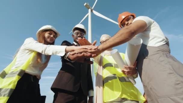 Skupina čtyř multikulturních partnerů drží ruce u sebe a usmívá se před kamerou, zatímco stojí mezi větrnými turbínami. Muži a ženy v bezpečnostních přilbách mají úspěšné setkání venku. — Stock video