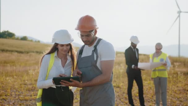 Cuatro colegas usando planos y tableta al aire libre. Colegas multirraciales controlando el trabajo de la granja de molinos. Eco para la energía eléctrica. Ingenieros exploran futuros proyectos de instalación de aerogeneradores. — Vídeo de stock