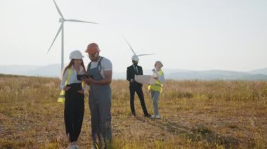 Uluslararası iş ortakları ekibi rüzgar türbinleri arasında bir arada duruyor ve planları inceliyor. Bir grup profesyonel, temiz çevre dostu enerji üretmek için işbirliği yapıyor. Eko elektrik gücü.